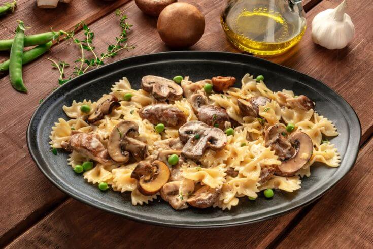 Pasta farfalle con funghi, pancetta e piselli, servita su un piatto nero di ceramica con dettagli di pancetta croccante e funghi freschi