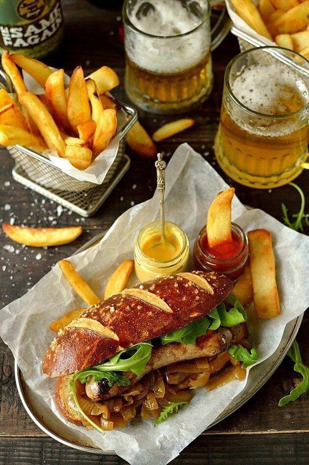 Un panino gourmet con wurstel grigliato, cipolle caramellate e insalata servito su un vassoio con carta da forno. Accanto ci sono patatine fritte spesse, due salse in vasetti di vetro (ketchup e senape) e due boccali di birra chiara con schiuma. Sullo sfondo, altri cestini di patatine e una bottiglia di birra.